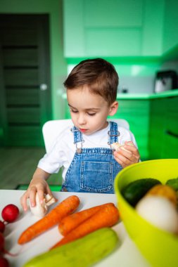 Çocuk mutfaktaki sebzeleri keşfediyor. Bir çocuk, etrafında havuç ve turpla merak göstererek, modern bir mutfak masasında taze sebzeleri inceliyor..