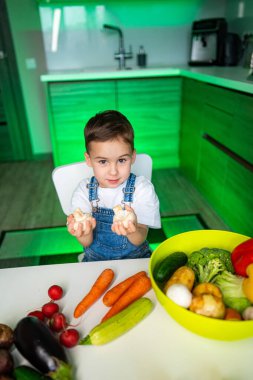 Çocuk mutfakta taze sebzelerin tadını çıkarıyor. Genç bir çocuk taze sebze masasına gülümsüyor, parlak bir mutfakta birkaç tane tutuyor..