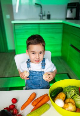 Çocuk mutfakta taze sebzelerin tadını çıkarıyor. Genç bir çocuk, modern bir mutfakta, renkli taze ürünlerle çevrili bir şekilde gülümserken sebzeleri tutuyor..