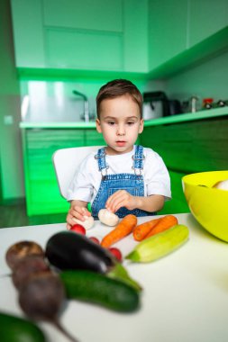 Çocuk mutfakta sebze ayırıyor. Tulumlu bir çocuk, modern bir mutfakta yeşil ışıkla dolu bir masada renkli sebzeler organize eder..