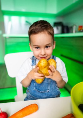 Küçük çocuk mutfakta patates tutuyor. Genç bir çocuk canlı, yeşil bir mutfakta patatesleri tutarken kot pantolon giyer ve gülümser..