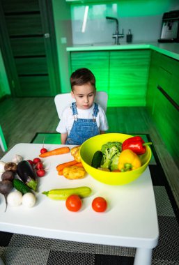 Çocuk evde taze sebzeleri keşfediyor. Renkli sebzeler ve meyvelerle çevrili bir masada oturan küçük bir çocuk sağlıklı yiyecek seçeneklerini araştırıyor..