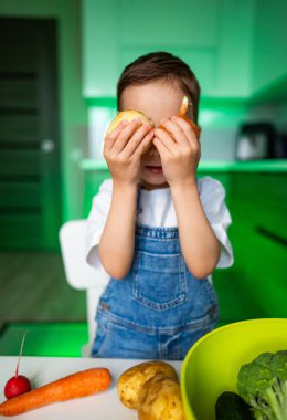 Çocuk mutfakta sebzelerle oynuyor. Genç bir çocuk yemek pişirmeye hazırlanırken parlak bir mutfakta gözlerinin önünde sebze tutuyor..