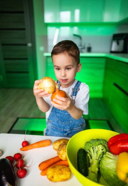 Mutfakta sebze arayan bir çocuk. Genç çocuk renkli ürünlerle uğraşırken elinde sebze tutuyor. Parlak bir mutfak ortamında..