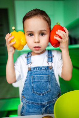 Mutfakta biberli çocuk. Kot pantolon giymiş genç bir çocuk parlak bir mutfakta sarı ve kırmızı biber gösterip sağlıklı beslenmeyi teşvik ediyor..
