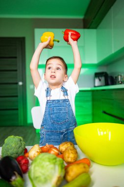 Çocuk sebzelerle yemek yapmayı seviyor. Genç çocuk, taze sebzeler ve karıştırma kabıyla çevrili modern bir mutfakta mutlu bir şekilde sarı ve kırmızı biber tutuyor..