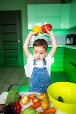 Çocuk sebzelerle yemek yapmaya bayılıyor! Genç bir çocuk mutfakta çeşitli taze sebzelerle çevriliyken sarı ve kırmızı dolmalık biber tutuyor..