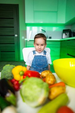 Çocuk taze sebzelerle gülümsüyor. Tulum giymiş neşeli bir çocuk renkli taze sebzelerle dolu bir mutfak masasında yemek yapmaya hazır..
