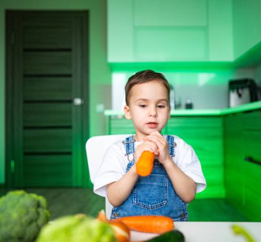 Çocuk modern mutfakta havuç tutuyor. Çağdaş bir mutfakta elinde havuçla ve çeşitli taze sebzelerle dolu bir masada oturuyor..