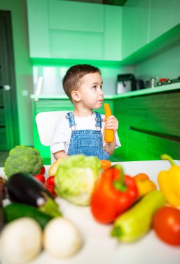 Çocuk mutfakta sağlıklı besleniyor. Parlak gözlü genç bir çocuk renkli sebzelerle dolu bir masada oturuyor ve elinde havuç tutuyor..