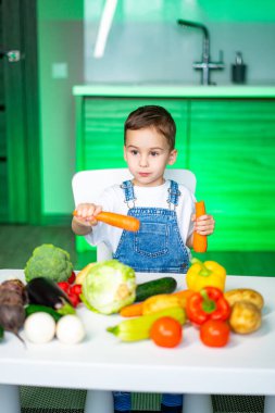 Küçük çocuk renkli sebzelerin tadını çıkarıyor. Kot pantolon giymiş bir çocuk havuç tutarken çeşitli taze sebzelerle çevrili, parlak bir mutfak ortamında..