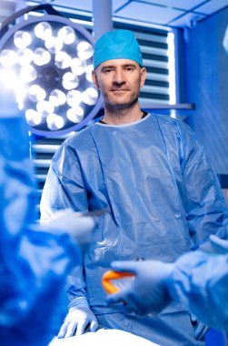 Cerrah ameliyata hazır. Meslektaşları ameliyathanede cerrahi aletleri hazırlarken mavi önlüklü bir cerrah güvenilir bir şekilde ayakta duruyor..