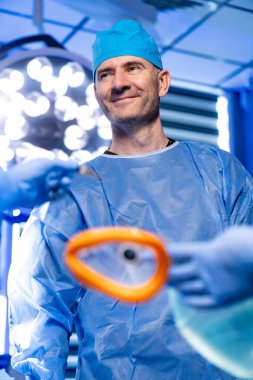 Cerrah ameliyata hazırlanıyor. Cerrah, iyi aydınlatılmış bir ameliyathanede ameliyat aletleri hazırlarken gülümseyerek ameliyat önlüğü ve eldiven giyer..