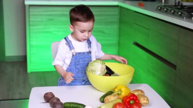 Çocuk mutfakta sebze doğruyor. Yeni yürümeye başlayan bir çocuk, parlak bir mutfakta renkli sebzeleri ayırıyor ve büyük bir kasede yemek pişirmeye odaklanıyor..