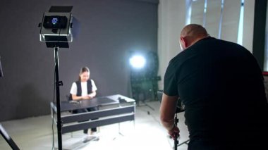 Siyah tişört giyen geniş omuzlu erkek fotoğrafçı kameraya arkasını döner. Kameraman, bulanık arka planda oturan bir bayanın görüntüleri için ekipmanı ayarlıyor..