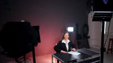 Beyaz gömlek ve siyah ceket giyen pozitif gülümseyen bayan fotoğraf stüdyosundaki masada oturuyor. Kadın muhabir kimliği. Çekimler için hazırlanıyor..