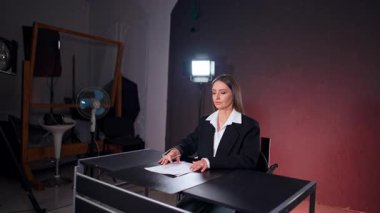 Siyah ceketli kadın muhabir fotoğraf stüdyosunda oturuyor. Sahne arkasında duran bir bayanın silueti..