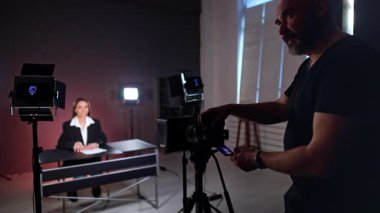 Erkek fotoğrafçı, masada oturan bir kadınla video kaydediyor. Kameraman kameraya bakar ve güler..