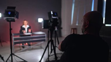 Sahne arkasında kameranın önünde oturan bir erkek fotoğrafçının arka görüntüsü. Siyah ceketli, tanınmayan uzun saçlı kadın masada oturmuş konuşuyor..