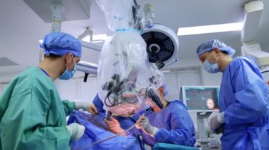 Tıp ekibindeki cerrahlardan biri mikroskobun arkasında plastikle kaplı oturuyor. Asistanı, bir tıbbi aleti beyin cerrahına verir..