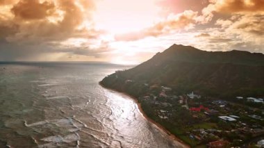 Batan güneş ışınları kümülüs bulutlarının arasından geçer. Ayaklarının dibinde resimli bir köyle güzel kayalar üzerinde uçuyor. Waikiki, Hawaii, ABD. Hava perspektifi.
