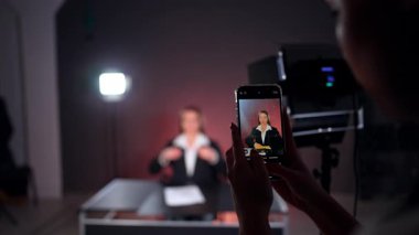 Telefon ekranında beyaz gömlek ve siyah ceket giyen uzun saçlı güzel beyaz bir bayan. Bayan muhabir stüdyoda çekim için hazırlanıyor. Bulanık arkaplan.