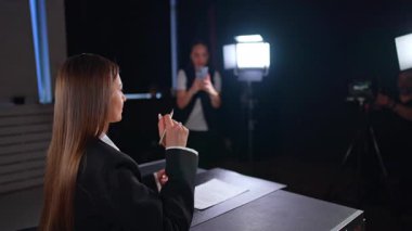 Siyah ceketli uzun saçlı esmer kadın çorapların ışığında oturuyor. Kameraman ve asistanının telefonuyla çekim yaptığı kız kuliste. Bulanık arkaplan.