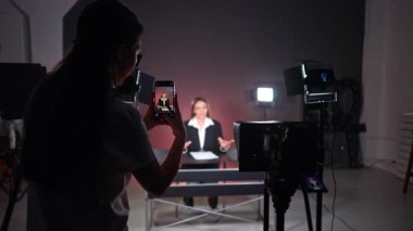 Telefonuyla fotoğraf çeken uzun saçlı bir esmerin arka görüntüsü. Kız sahne arkasındaki video ekipmanlarının arasında duruyor..