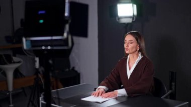 Uzun kahverengi saçlı çekici beyaz kadın stüdyoda masa başında oturuyor. Olumlu bayan gülümsüyor.