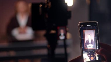 Erkek eller akıllı telefon tutarak kamera görüntülerini çekiyor. Kapatın. Kamera tripoda, bayan da bulanık fonda..