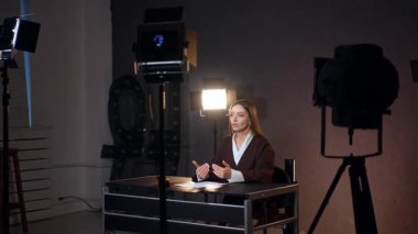 Duygusal beyaz kadın modern fotoğraf stüdyosunda kameraya güvenle konuşuyor. Soffits muhabirin etrafında duruyor..