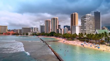 Dalgalar Waikiki, Honolulu, Hawaii, ABD 'deki kumlu plajları koruyan barajlara doğru hareket ediyor. İnsanlar Pasifik Okyanusu kıyısında dinleniyor. Gökyüzünü kaplayan gri bulutlar manzaranın üzerinde.