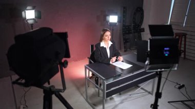 Siyah ceket giyen beyaz kadın stüdyodaki masada oturuyor. Kadın, çorapların ışığında kameraya konuşuyor. Yüksek açı görünümü.
