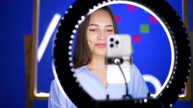 Gülümseyen beyaz kadın, telefonun arkasında üç ayaklı ve hafif yüzüklü duruyor. Video çeken pozitif blog yazarı.