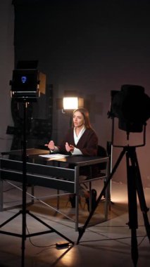 Modern stüdyoda blog çekimlerinin kulisinde. Esmer kadın masa başında oturmuş blog yazarken el kol hareketi yapıyor. Dikey video.