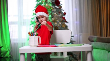 Noel Baba şapkalı bebek kulağına telefon tutuyordu. Çizmek için kalem seçen sevimli çocuk. Arkaplanda Noel ağacı.