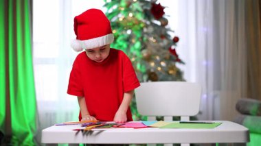 Kırmızı Noel Baba şapkalı ve tişörtlü sevimli bebek odasındaki masada duruyor. Küçük çocuk bir kalem seçiyor ve çizmeye başlıyor. Bulanık arkaplanda Noel ağacı.