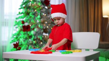 Kırmızı Noel Baba şapkalı ciddi bir çocuk masada oynuyor. Sevimli çocuk evde bulmaca çözüyor. Arkaplanda Noel ağacı.