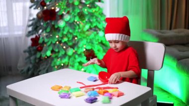Kırmızı Noel Baba şapkalı sevimli çocuk makası kullanarak plastik parçaları kesiyor. Çocuk Noel için evde sanat hobisi yapıyor..