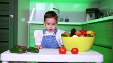 Kot pantolon giyen küçük beyaz çocuk mutfaktaki masada oturuyor. Bebek bir kase sebzeyi yakına çeker ve bir patlıcan seçer..