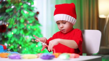 Masada, Noel Baba şapkası takan konsantre beyaz çocuk oynuyor. Sevimli erkek bebek makas ve plastikle oynuyor. Bulanık arkaplanda Noel ağacı.