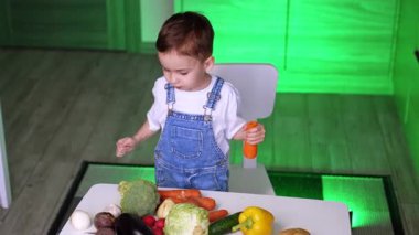 Beyaz tişörtlü ve kot pantolonlu yakışıklı bebek mutfaktaki masada duruyor. Havuç tutan çocuk önündeki çeşitli sebzelere bakıyor. Yüksek açı görünümü.