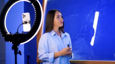 Uzun saçlı kadın cam duvarda duruyor ve beyaz kalemle yazıyor. Kadın blogcu ışık halkalı bir telefon kamerasıyla yayın yapıyor.