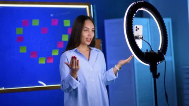 Genç bir kadın çevrimiçi içerik yaratır. Genç bir kadın, canlı yayın ya da video için halka ışığının önünde dururken enerjik bir şekilde konuşur..