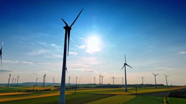 Güneşli bir arazide rüzgar türbinleri. Uçsuz bucaksız yeşil bir alanda, dağınık bulutlu, açık mavi bir gökyüzünün altında birkaç rüzgar türbini dönüyor..