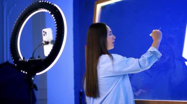 Stüdyoda sanat eseri yaratan genç bir kadın. Genç bir kadın, sanat malzemeleri ve ekipmanlarıyla çevrili, iyi ışıklandırılmış bir stüdyonun içindeki mavi bir tuvale yaratıcı bir şekilde yazar..