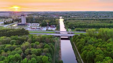 Kanal gün batımı ve yeşillik. Güzel bir günbatımı, ağaçlarla çevrili bir kanalın üzerine sıcak renkler serer..