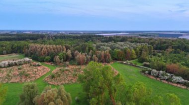 Huzur dolu yeşil alanlar. Geniş bir arazide alacakaranlık sırasında sakin bir gökyüzü altında çeşitli ağaçlar, tarlalar ve uzak binalar bulunur..