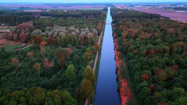 Kanal kenarında sonbahar ağaçları. Berrak mavi gökyüzü altında canlı sonbahar renklerini gösteren ağaçlarla çevrili sakin bir kanalın havadan görünüşü.