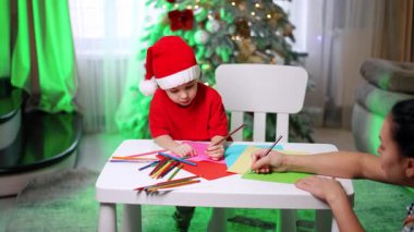 Çocuk ailesiyle tatil sanatı yaratıyor. Noel şapkalı küçük bir çocuk kağıdın üzerinde renklendirilirken, ebeveyn yardımcı oluyor, şenlikli dekorasyonlarla çevrili..